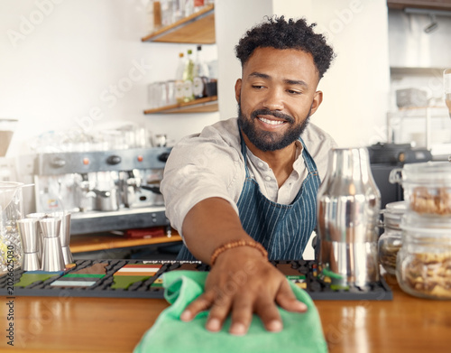 Tableau sur toile Waiter, man and cleaning in coffee shop with cloth, hospitality hygiene and disinfection for bacteria