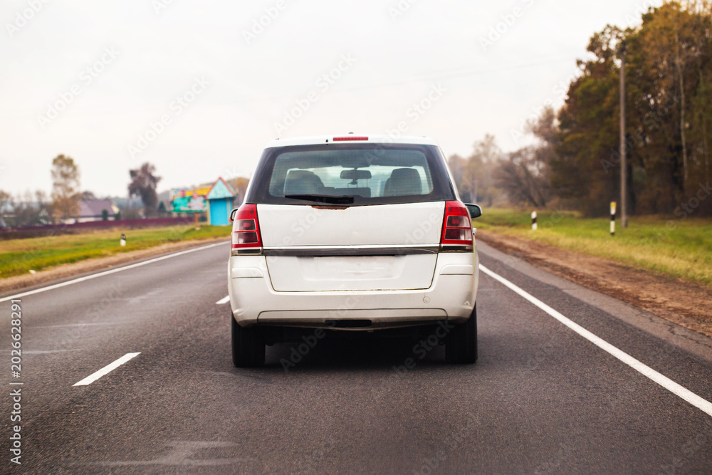Fototapeta premium White color family car minivan or station wagon traveling on asphalt road through rural landscape under overcast sky daytime autumn season