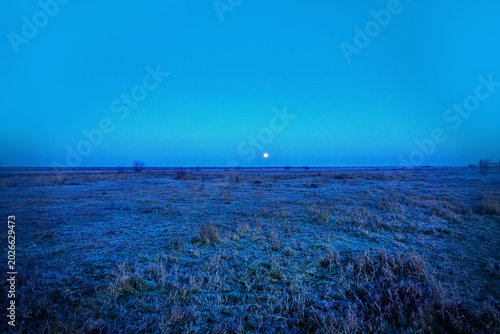 Astronomy. A rare phenomenon of the morning full-orbed moon setting covered by a light mist, swamp miasma, civil twilight. Spring sunrise over desert weadow