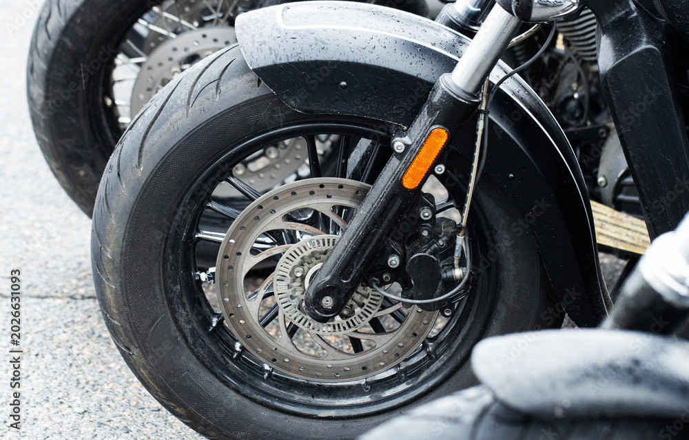 Obraz premium Close-up view of front wheel of motorcycle, showing tire, brake disc, suspension, and fender covered in raindrops, vehicle parked outdoors on asphalt, detail of modern bike engineering