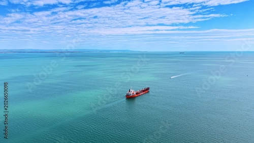 Industrial oil tanker vessel cruising turquoise ocean water. High angle drone shot of a red tanker ship moving through calm turquoise sea with distant coast.