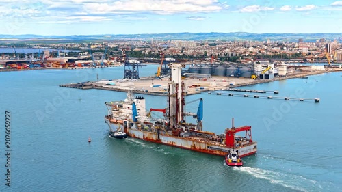 Marine specialized drilling ship navigating near industrial port terminal. Technical aerial drone view of offshore well intervention vessel accompanied by tugboat at commercial seaport.