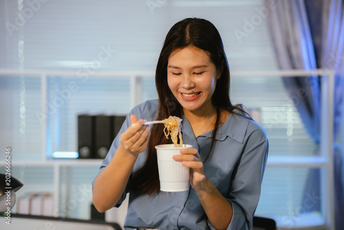 Young Asian woman eating instant noodles while working late at office desk with tablet and computer, representing overtime work and busy lifestyle.