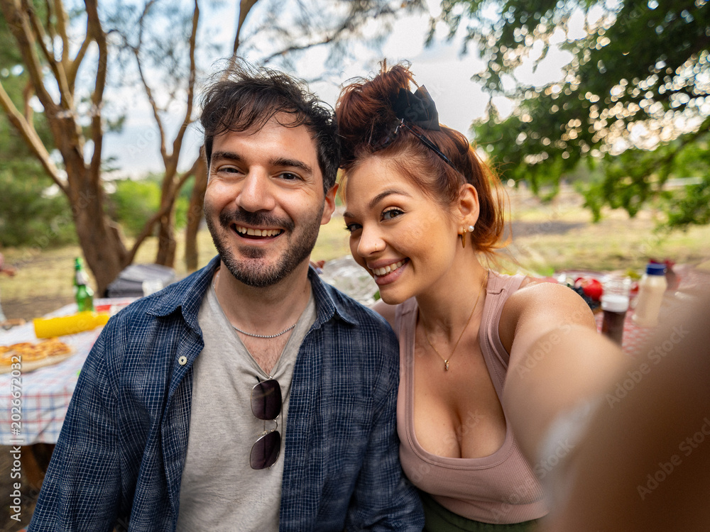 Fototapeta premium Happy young couple smiling while taking selfie during outdoor picnic in park