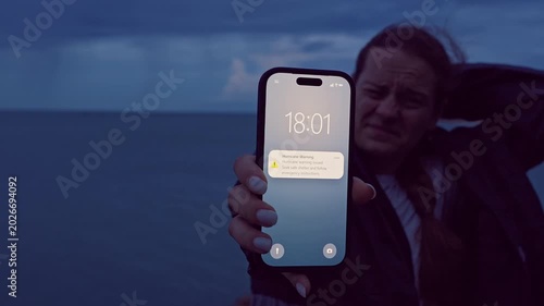 Woman Holding Smartphone With Hurricane Warning While Struggling Against Strong Coastal Wind at Dusk, Emergency Alert and Severe Weather Survival