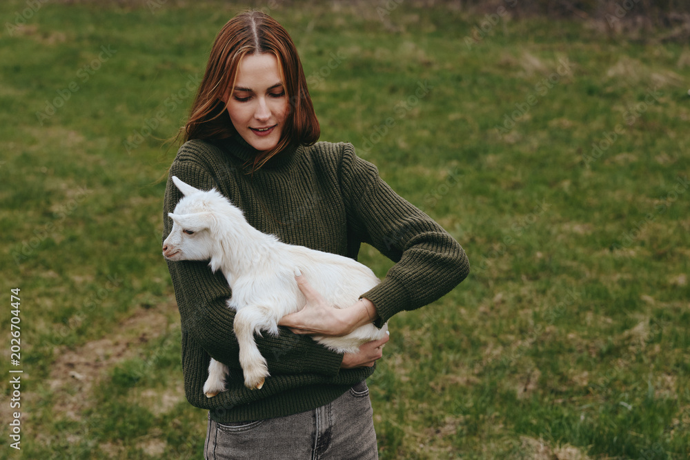Fototapeta premium Lifestyle scene of a woman in a knitted green sweater holding a goat kid in a field, meadow and nature backdrop, warm light, calm affectionate moment outdoors.