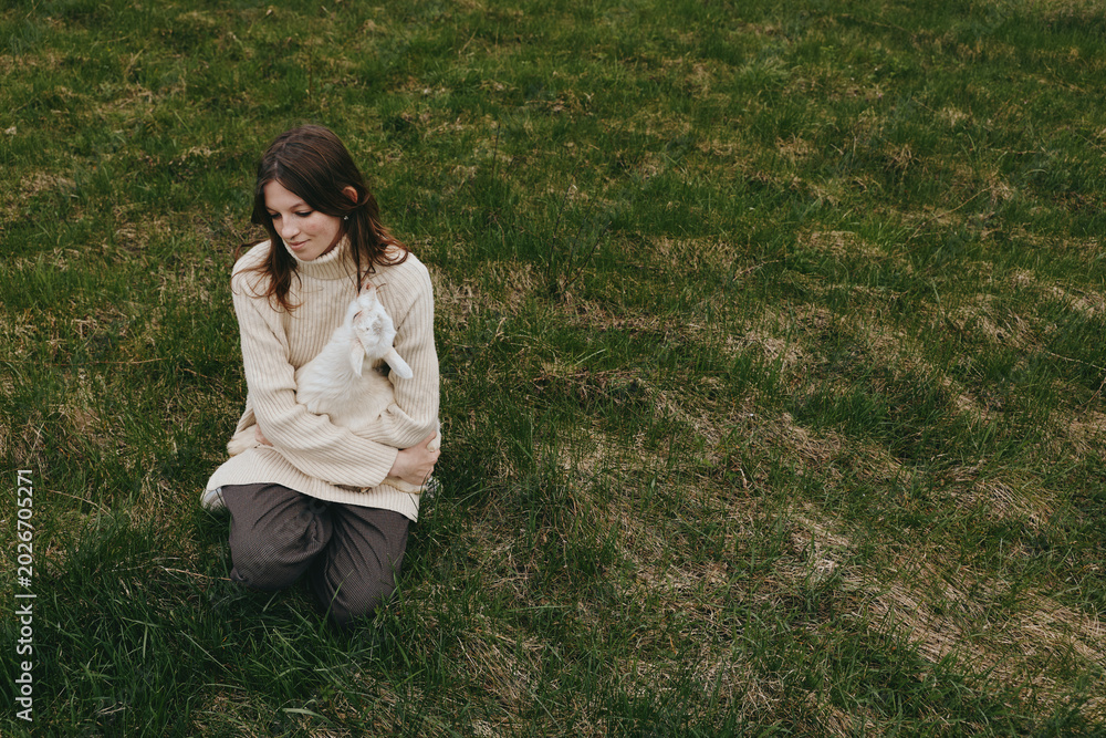 Fototapeta premium A casual lifestyle scene shows a young woman kneeling on grassy ground, cuddling a small white animal, cozy sweater, tranquil moment outdoors, conveying tenderness and connection with pets.