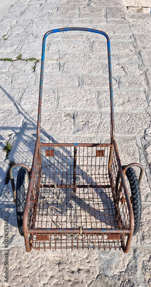 Obraz premium High angle view of an old weathered rusty metal wire cart with two wheels standing on a stone paved pier.