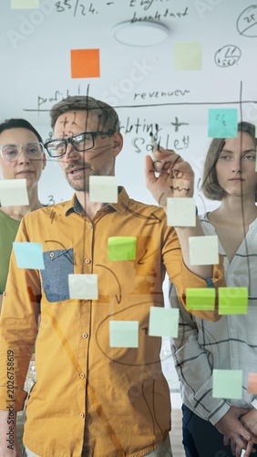 Business people team brainstorming, sharing strategy on glass whiteboard with colorful sticky notes, working collaboratively in bright office