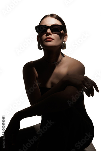 Woman in black sunglasses posing in dramatic shadow with elegant jewelry. Concept of eyewear fashion ad, luxury accessory campaign, bold beauty editorial and modern style branding.