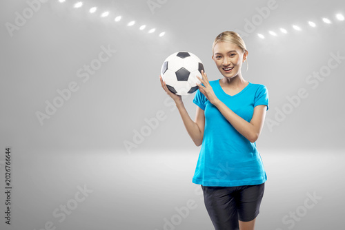A joyful young Indonesian southeast asian woman with a bright smile holds a soccer ball against a blurred stadium background with glowing lights. Perfect for sports and fitness themes