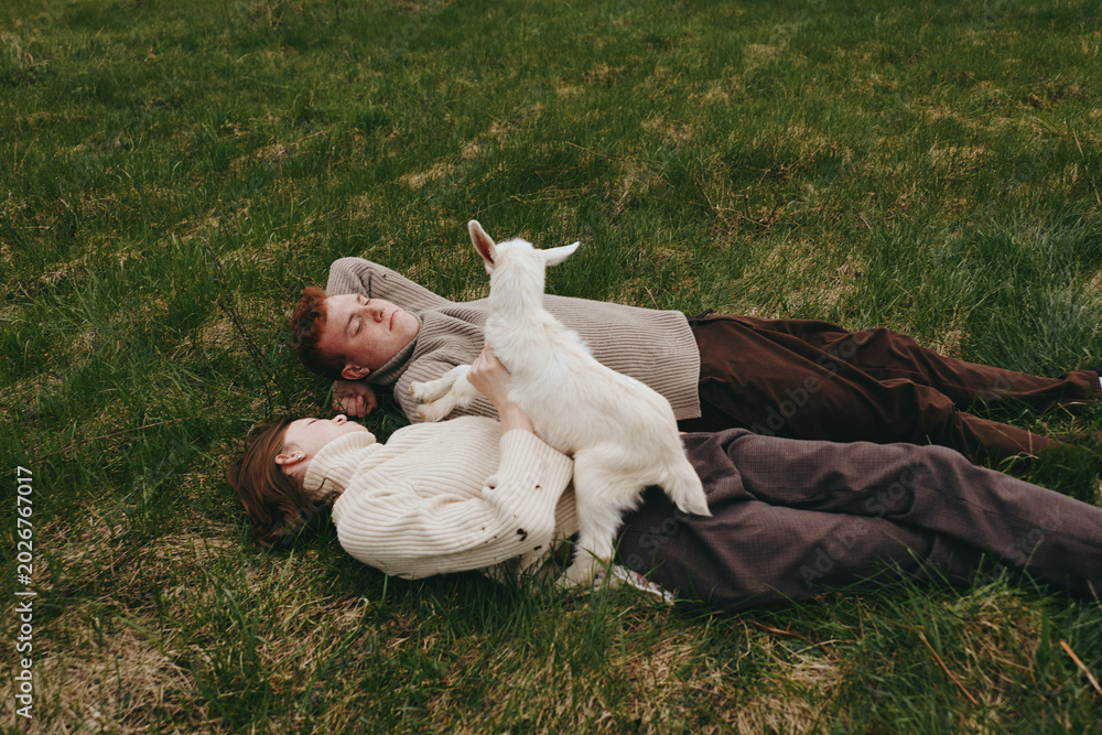 Fototapeta premium Relaxed couple lying on grass with a curious goat kid climbing nearby. Warm earthy tones highlight outdoor lifestyle, companionship, and gentle play. Soft daylight creates a cozy, candid moment of