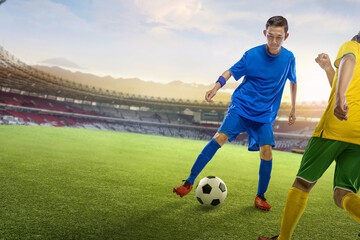 Professional Indonesian southeast asian football player in blue uniform controls the ball during a match at a packed stadium under a sunny sky with mountains in the background