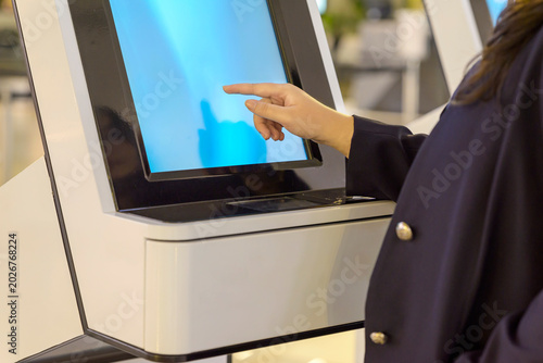 Close-up of a person's hand interacting with a digital touch screen on a modern white self-service kiosk, likely for check-in or service in a public place