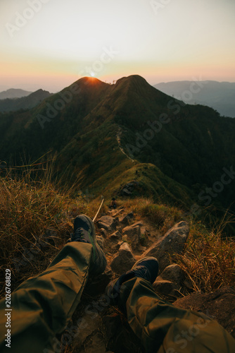 The frame shows the hiker's legs in green trekking pants and sturdy hiking boots resting on rugged rocks and dry grass in the foreground. A narrow,