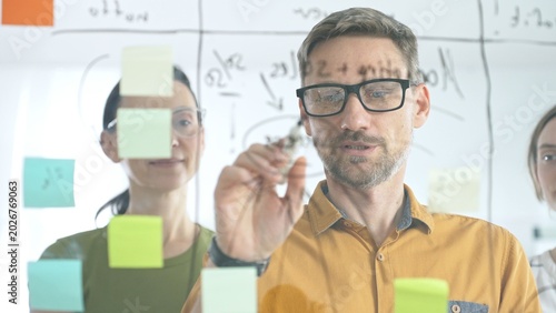 Professional team brainstorming project strategy, placing colorful sticky notes on glass board, analyzing data, sharing insights, developing collaborative solutions