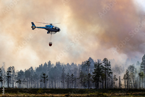 Helicopter flying with water bucket in smoky sky above forest
