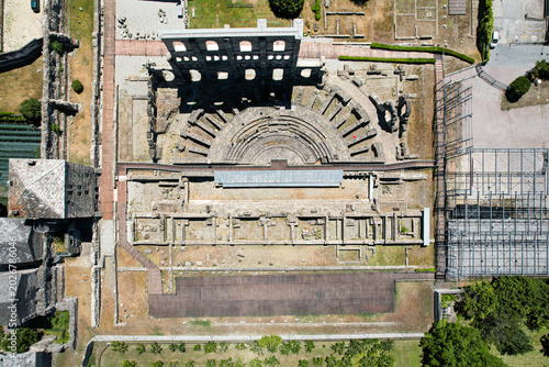 Aerial view of the Roman Theatre of Aosta featuring the semicircular stone seating and the high arched facade wall among ancient ruins in Aosta, Aosta Valley, Italy.