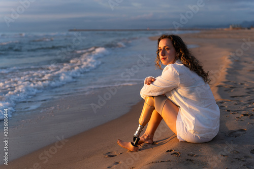 Woman with a prosthetic leg sitting on the beach at sunset reflecting calmly