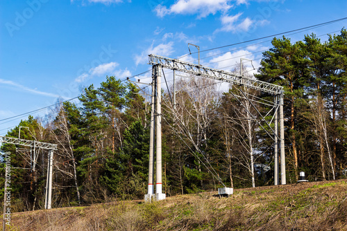 Industrial steel railway overhead line structures and electric power wires towering over the green forest edge against a bright blue sky on a sunny day in the countryside