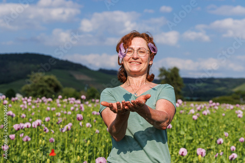Mature woman with glasses and curly red hair playfully throwing pink poppy petals while standing in a blooming poppy field.