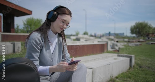A hand-held shot of a young female student in casual clothes, wearing glasses and headphones, sitting on a concrete in an urban park on a sunny summer day and listening to music from a mobile phone