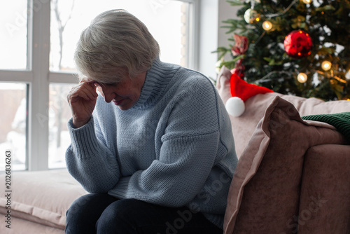 Fotografía Sad elderly woman sitting on sofa near Christmas tree