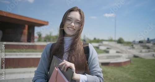 A hand-held portrait of a young female student in casual clothes, wearing glasses and a backpack, holding a notebook and laptop in her hands and posing looking at the camera while standing in a park 