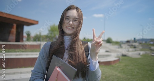 A hand-held portrait of a young female student in casual clothes, wearing glasses and a backpack, holding a notebook and laptop in her hands and making a welcoming gesture with her hands - victory