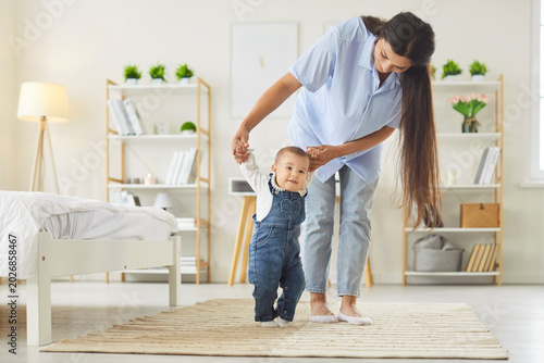 Foto Happy young mother helping her cute little baby with first steps at home