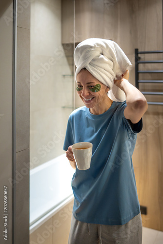 Konstfotografi Happy smiling woman with towel on head, eye patches holding mug, quickly getting ready in bathroom before work or daily routine