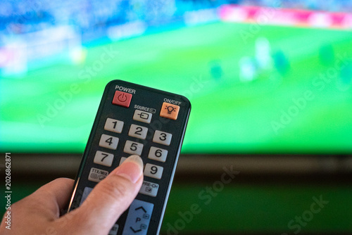 Hand Holding Remote Control Pointing at Television showing football match in action.