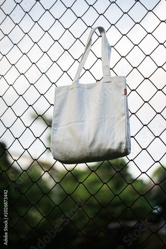 White tote bag hangs on chain-link fence. Soft lighting highlights fabric texture and simple design. Background shows grassy field and blurred trees. Casual