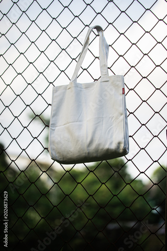 White tote bag hangs on chain-link fence. Soft lighting highlights fabric texture and simple design. Background shows grassy field and blurred trees. Casual
