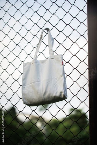 White tote bag hangs on chain-link fence. Soft sunlight highlights fabric's gentle folds. Background features blurred green grass and distant trees. No people visible, suggesting quiet, empty scene