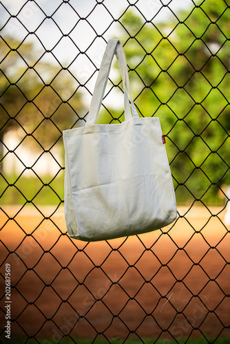 Simple cotton tote hangs on chain-link fence. Sunlight glows behind green trees and grass. Tote shows soft folds and clean lines. Natural scene suggests casual, everyday use