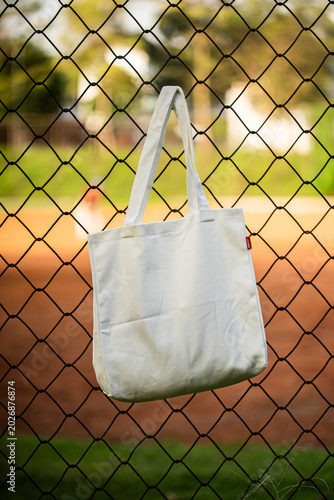 White tote bag hangs on chain-link fence. Blurry background shows green grass and dirt field. Child in distance wears cap and holds bat. Sunlit scene suggests casual park or baseball setting