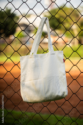 Simple tote bag hangs on chain-link fence. Soft fabric shows subtle wear and natural folds. Background holds green trees and blurred backyard. Scene suggests casual, everyday outdoor setting