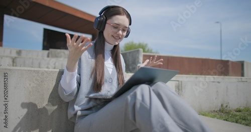 Photos A hand-held shot of a young female student in casual clothes, glasses, and headphones making a video call on a laptop while sitting on a concrete bench in an urban park