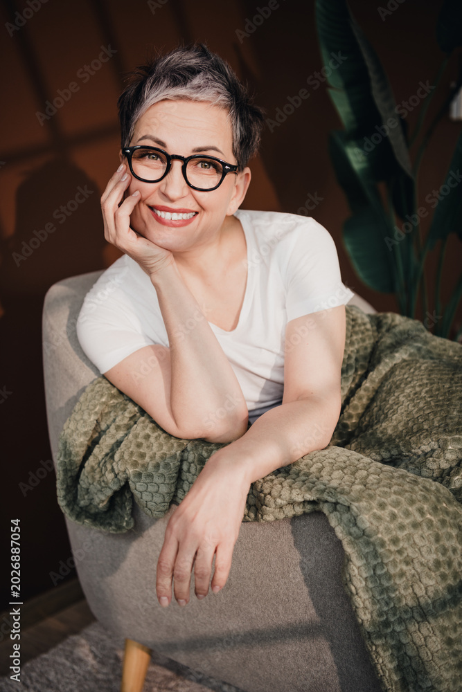 Fototapeta premium Smiling mature woman with glasses relaxing in an armchair wrapped in a green blanket, cozy home portrait conveying warmth, confidence and contentment