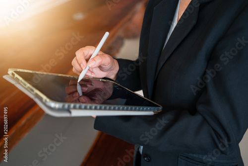 Side view close-up of a businesswoman hands using a stylus pen to sign a contract, draw, or tap on a digital tablet screen at a wooden desk, with soft morning sunlight.