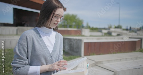 Fotografía A hand-held shot of a young female student in casual clothes and glasses reading a book while flipping through pages while sitting in an urban park