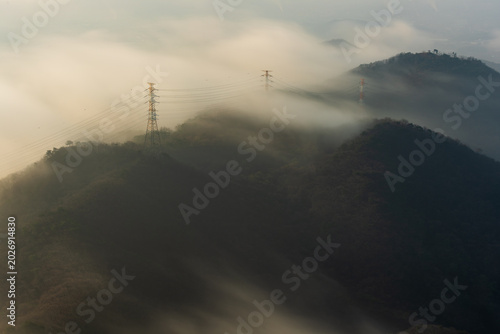 Schilderij op canvas Amazing sea of clouds flowing like a waterfall over a mountain ridge with electricity pylons during a beautiful pastel sunrise