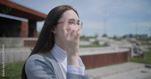 A handheld shot of a young female student in casual clothes sitting on a concrete bench in an urban park, enjoying a sunny summer day.