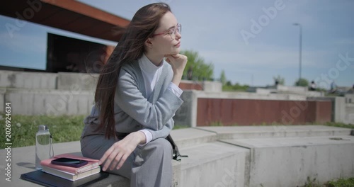 A handheld shot of a young female student in casual clothes sitting on a concrete bench in an urban park, enjoying a sunny summer day.
