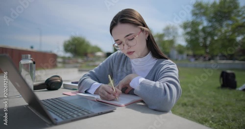 A hand-held shot of a young female student in casual clothes and glasses using a laptop and taking notes with a pen in a notebook in an urban park.