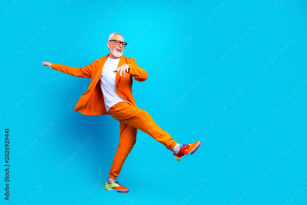 Fototapeta premium Joyful senior man dancing in bright orange suit against a blue background, stylish retiree celebrating life with energetic pose and colorful sneakers