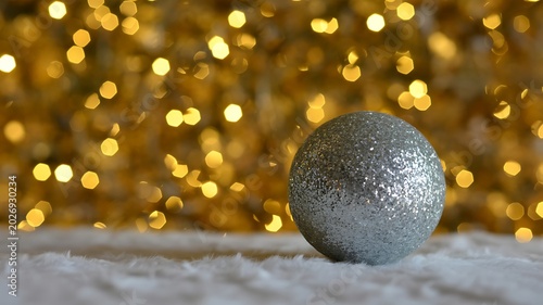 Silver glitter christmas ornament resting on white fur with soft focus bokeh golden lights in the background for festive holiday celebration