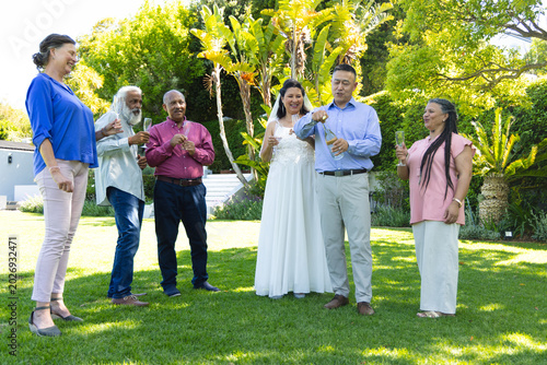 Groom popping champagne, bride wearing wedding gown holding flute, guests celebrating on lawn
