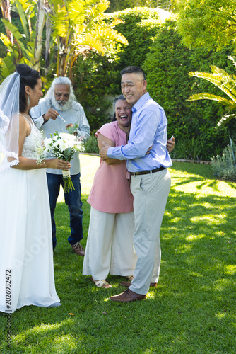 Diverse family gathering on lawn with couple hugging, bride in gown and veil holding bouquet